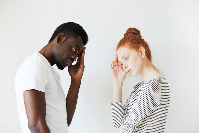 Sideways portrait of couple in disappointed pose in white studio. Caucasian girl resting her head on her hand as if thinking and solving the problem, African man also looking puzzled and stressed.